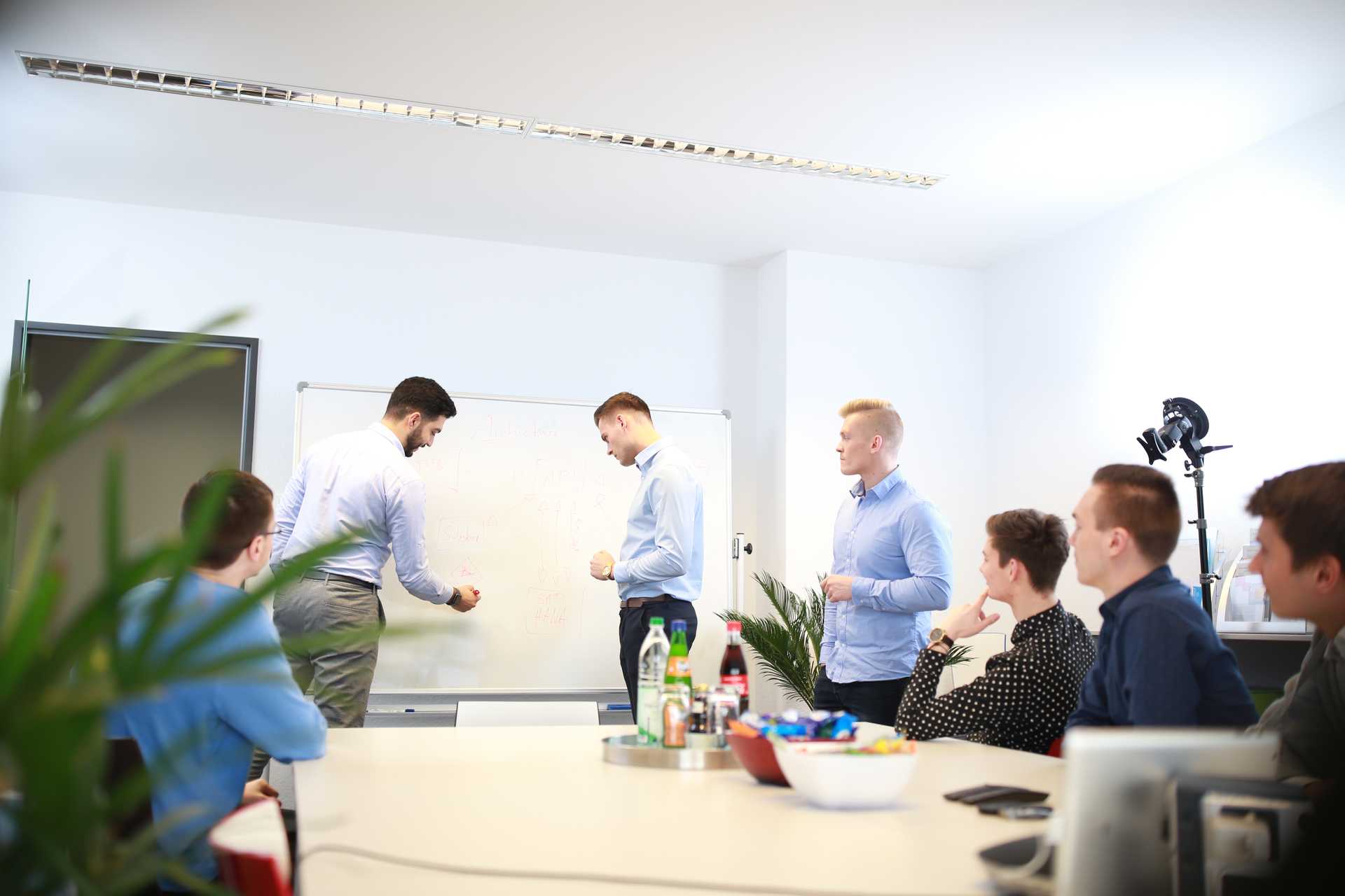 Das Incoqnito Team sitzt gemeinsam in einem Besprächungsraum. Zwei Mitarbeiter stehen und präsentieren vor einem Whiteboard.