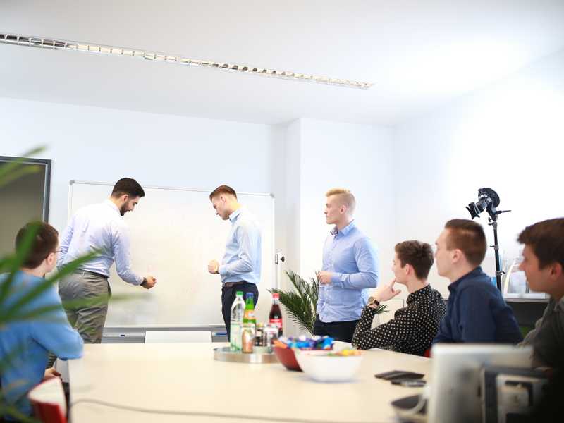 Das Incoqnito Team sitzt gemeinsam in einem Besprächungsraum. Zwei Mitarbeiter stehen und präsentieren vor einem Whiteboard.